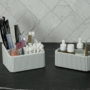 May include: Two matching gray ribbed cosmetic organizers on a black marble surface. One organizer holds makeup brushes, mascara, and cotton swabs. The other holds skincare products and a pencil. The background features a white marble tile wall.