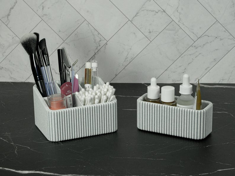 May include: Two matching gray ribbed cosmetic organizers on a black marble surface. One organizer holds makeup brushes, mascara, and cotton swabs. The other holds skincare products and a pencil. The background features a white marble tile wall.