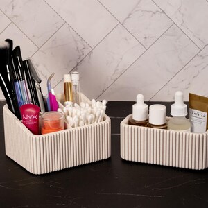 May include: Two off-white, ribbed cosmetic organizers on a black surface. One holds makeup brushes, applicators, and products, including a tube of NYX lipstick. The other contains skincare products. The background is a white marble-patterned wall.