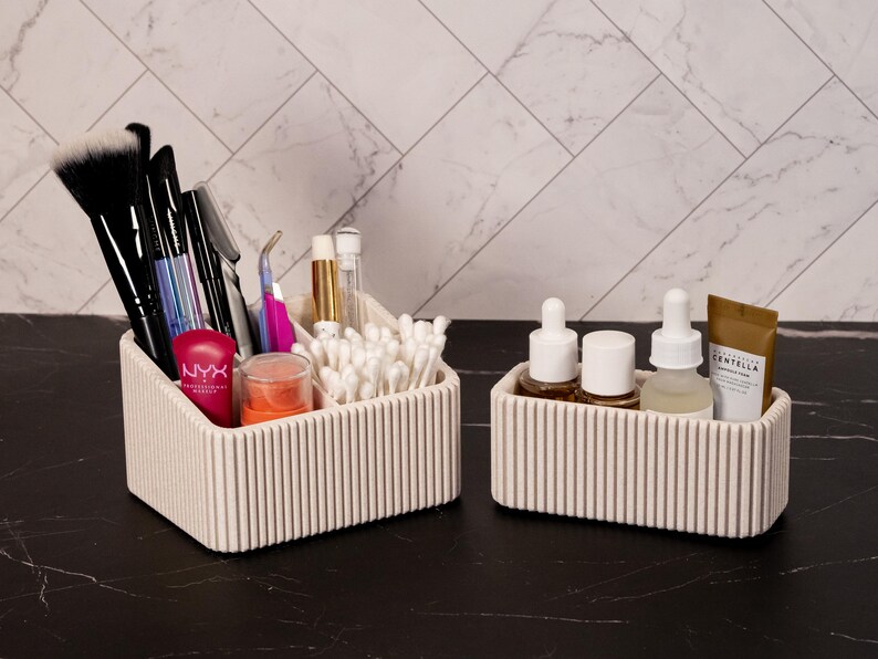May include: Two off-white, ribbed cosmetic organizers on a black surface. One holds makeup brushes, applicators, and products, including a tube of NYX lipstick. The other contains skincare products. The background is a white marble-patterned wall.