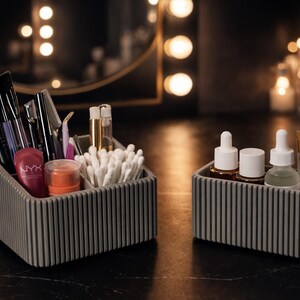 May include: Two gray, ribbed, rectangular organizers on a dark surface. One contains makeup brushes, applicators, and products, including a tube of NYX lip product. The other holds skincare bottles and a small tube of Centella product.