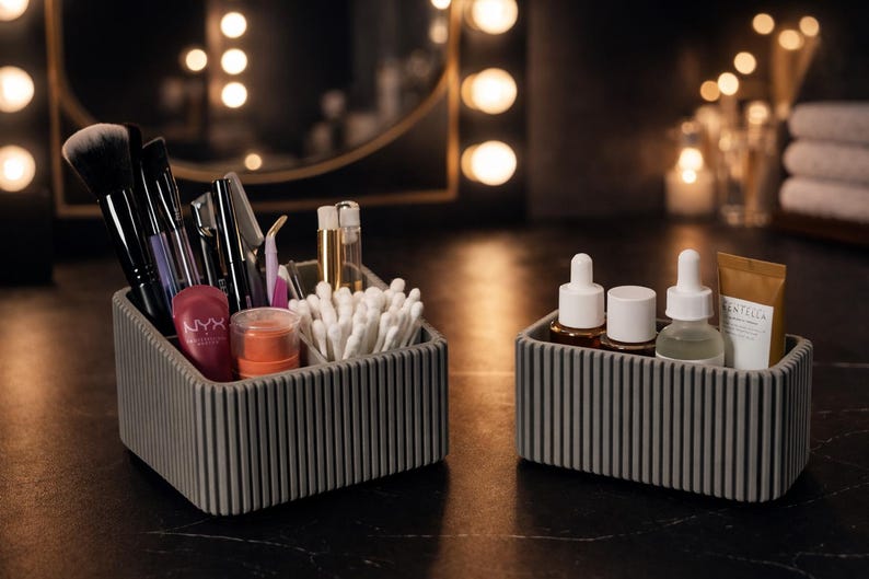 May include: Two gray, ribbed, rectangular organizers on a dark surface. One contains makeup brushes, applicators, and products, including a tube of NYX lip product. The other holds skincare bottles and a small tube of Centella product.
