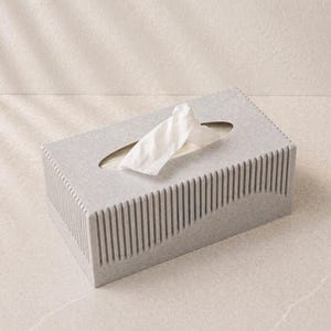 May include: A rectangular tissue box in a light gray color with vertical lines on the sides and a curved design. White tissues are pulled from the oval opening on top. The box is on a light-colored surface.
