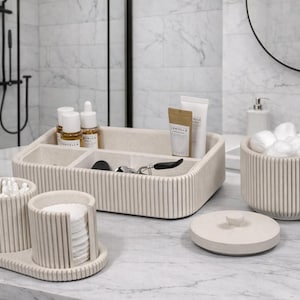 Puede incluir: Un conjunto de organizadores de baño beige, que incluye una bandeja con compartimentos, un tarro para bolas de algodón y recipientes para bastoncillos y discos de algodón. Los artículos tienen un diseño acanalado y están colocados sobre una encimera de mármol.