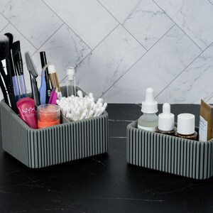 May include: Two gray, ribbed cosmetic organizers on a black surface. The larger organizer holds makeup brushes, pens, and cotton swabs. The smaller organizer contains skincare products, including bottles and tubes. The background features a white and gray tiled wall.