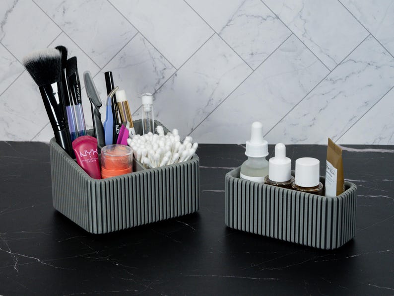 May include: Two gray, ribbed cosmetic organizers on a black surface. The larger organizer holds makeup brushes, pens, and cotton swabs. The smaller organizer contains skincare products, including bottles and tubes. The background features a white and gray tiled wall.