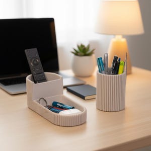 May include: A beige desk organizer with vertical ribbed detailing. It features compartments for a remote control, keys, batteries, and a separate cylindrical pen holder filled with pens and markers. A laptop and lamp are in the background.