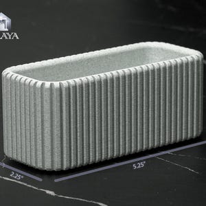 May include: A rectangular, light gray planter with vertical ribbed detailing. The planter measures 5.25 inches long and 2.25 inches wide and tall. The logo "VALAYA" is in the upper left corner.