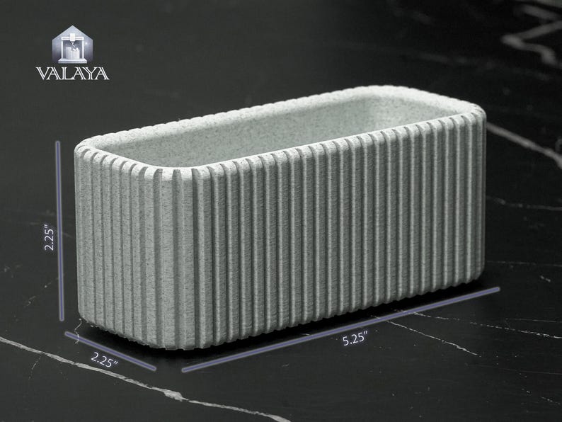 May include: A rectangular, light gray planter with vertical ribbed detailing. The planter measures 5.25 inches long and 2.25 inches wide and tall. The logo "VALAYA" is in the upper left corner.