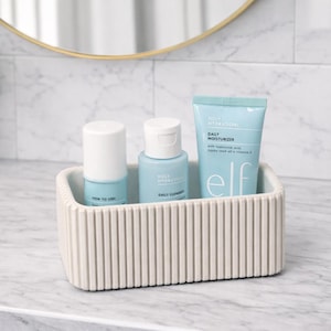 May include: A light beige, ribbed rectangular container holds three blue and white skincare products. The products include a roller, a bottle, and a tube of Daily Moisturizer. The products are labelled with the brand name "elf". The container sits on a marble countertop.