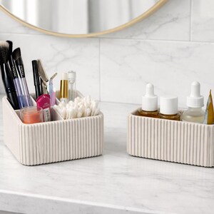 May include: Two off-white, ribbed cosmetic organizers on a marble countertop. One organizer holds makeup brushes, pens, and cotton swabs. The other holds skincare bottles and a tube of product. A small potted plant and a mirror are in the background.