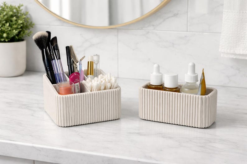 May include: Two off-white, ribbed cosmetic organizers on a marble countertop. One organizer holds makeup brushes, pens, and cotton swabs. The other holds skincare bottles and a tube of product. A small potted plant and a mirror are in the background.