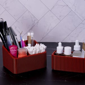 May include: Two red wooden organizers, one larger and one smaller, on a black surface. The larger organizer holds makeup brushes, cotton swabs, and cosmetic products. The smaller organizer contains skincare bottles and tubes. The background features a white marble pattern.