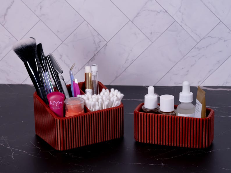 May include: Two red wooden organizers, one larger and one smaller, on a black surface. The larger organizer holds makeup brushes, cotton swabs, and cosmetic products. The smaller organizer contains skincare bottles and tubes. The background features a white marble pattern.