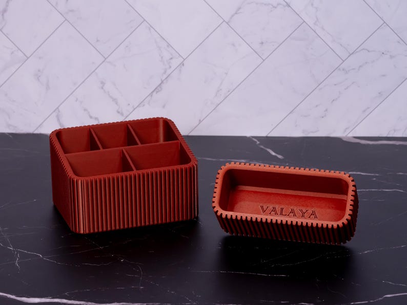 May include: Two terracotta-colored organizers. One is rectangular with six compartments, the other is a smaller rectangular tray. The word "VALAYA" is etched on the smaller tray. Both have a ribbed design.