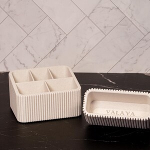 May include: Two beige organizers on a black surface. One is a rectangular container with six compartments. The other is a smaller, rectangular tray with the word "VALAYA" etched on the inside. Both have vertical ribbed detailing.
