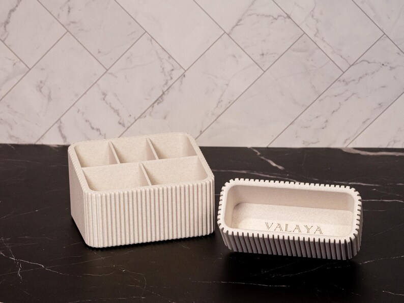May include: Two beige organizers on a black surface. One is a rectangular container with six compartments. The other is a smaller, rectangular tray with the word "VALAYA" etched on the inside. Both have vertical ribbed detailing.