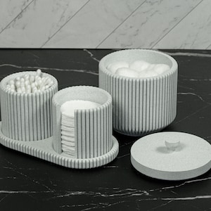 May include: A set of three light gray bathroom organizers. One container holds cotton balls, another holds cotton swabs, and a third holds cotton pads. A matching lid sits nearby. The organizers have a vertical striped design.
