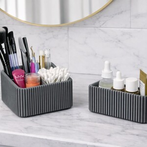 May include: Two gray, ribbed cosmetic organizers on a marble countertop. One organizer holds makeup brushes, applicators, and beauty products. The other holds skincare bottles and a small tube. A mirror and plant are in the background.
