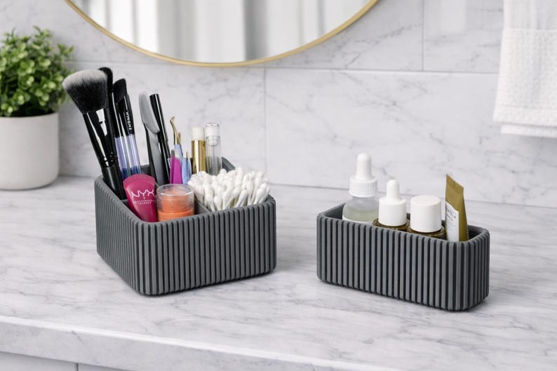 May include: Two gray, ribbed cosmetic organizers on a marble countertop. One organizer holds makeup brushes, applicators, and beauty products. The other holds skincare bottles and a small tube. A mirror and plant are in the background.