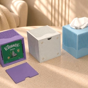 Puede incluir: Tres fundas cuadradas para cajas de pañuelos con texturas acanaladas verticales en morado, blanco y azul claro. La funda morada está abierta, revelando una caja de pañuelos Kleenex Lotion verde dentro, con su tapa morada a juego delante.