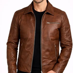 Può includere: Giacca in pelle marrone con chiusura a zip e collo a camicia. La giacca presenta un design classico con una tasca sul petto e due tasche laterali. Il modello indossa una maglietta nera sotto.