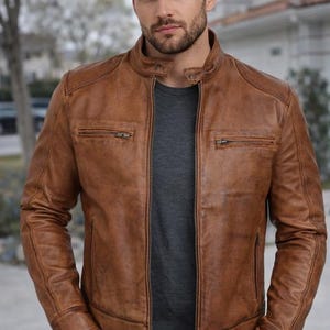 May include: A brown leather jacket with a stand-up collar and two zippered chest pockets. The jacket has a full-length zipper closure and is worn over a gray t-shirt. The jacket is a medium brown color.