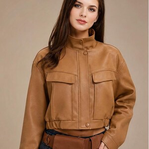 Könnte beinhalten: Eine braune, kurze Lederjacke mit zwei Vordertaschen und einem hohen Kragen. Die Jacke wird mit einer braunen Bauchtasche und einer blauen Jeans kombiniert. Die Jacke hat einen Knopfverschluss.