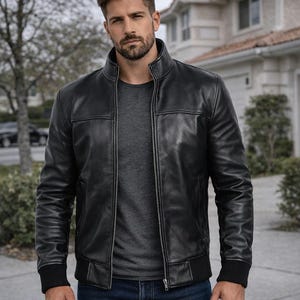 May include: A black leather bomber jacket with a zip-up front, ribbed cuffs, and a stand-up collar. The jacket has a classic design with visible stitching details. The model is wearing a gray shirt and blue jeans.