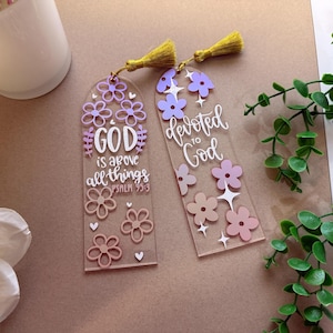 May include: Two clear acrylic bookmarks with floral designs and gold tassels. One bookmark reads "GOD is above all things" with Psalm 95:3. The other bookmark reads "devoted to God". The flowers are shades of pink and purple.