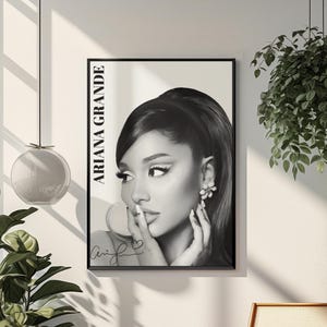 May include: Black and white framed poster featuring Ariana Grande. The poster has the text "ARIANA GRANDE" vertically on the left side. The image shows a close-up of Ariana Grande with her finger on her lips. The poster is hanging on a white wall.