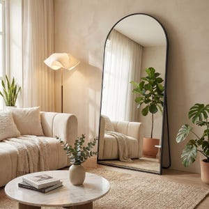 May include: A large, arched-top floor mirror with a black metal frame stands in a living room. The mirror reflects a cozy seating area with a beige sofa, a round coffee table, and potted plants. The room has a neutral color palette.