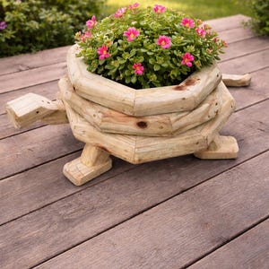 May include: A wooden turtle-shaped planter filled with green foliage and vibrant pink flowers. The planter is constructed from light-coloured wood and features a detailed design, including a head, tail, and four legs. The planter is placed on a wooden deck.