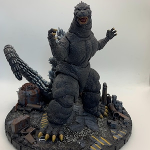 Könnte beinhalten: Eine dunkelgraue Godzilla-Actionfigur mit strukturiertem, schuppigem Körper, scharfen Krallen und einem langen Schwanz. Die Figur steht auf einem runden Sockel, der eine detaillierte, mit Trümmern übersäte Stadtlandschaft mit rostigen Metallelementen und gelben Warnstreifen zeigt.