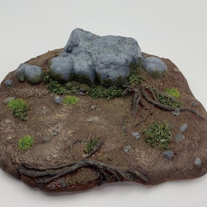 Könnte beinhalten: Eine Miniatur-Geländebasis mit einer großen grauen Felsformation, grünem Moos und kleinen Felsen. Die Basis ist braun mit detaillierter Textur, kleinen grünen Pflanzen und freiliegenden Wurzeln.