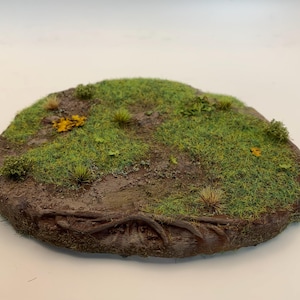 Op de afbeelding: Een ronde miniatuurterreinbasis met een gedetailleerd landschap. Het heeft groen gras, kleine planten en stukken aarde. De randen zijn donkerbruin en getextureerd om op boomwortels te lijken.