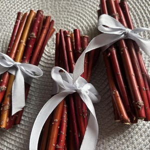 May include: Three bundles of red and orange twigs tied with white satin ribbon. The twigs have a natural, rustic appearance, with visible bark and varying shades of red and orange. The ribbon is tied in neat bows, adding a touch of elegance to the natural elements.