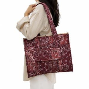 Peut inclure: Un sac fourre-tout matelassé arborant un imprimé bandana cachemire bordeaux foncé et blanc. Le sac présente une forme rectangulaire, deux bandoulières et une poche avant. Le tissu possède une texture piquée créant un aspect rembourré.