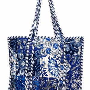 May include: A quilted tote bag featuring a patchwork design of various blue and white floral patterns. The bag has two long shoulder straps and edges finished with a blue and white striped fabric trim, creating a consistent visual texture.