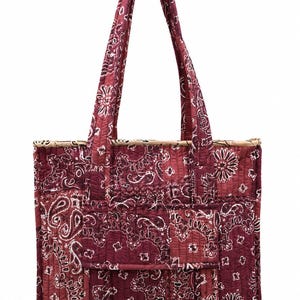 Op de afbeelding: Een gewatteerde tote bag met een donkerbordeaux en witte paisleyprint. De tas heeft twee lange schouderbanden, een rechthoekig lichaam met verticale stiksels en een voorvak met een klepsluiting. De bovenrand is afgewerkt met een beige bies.