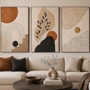 May include: Three framed abstract art prints with neutral tones and black accents hang above a cream-colored sofa. The artwork features geometric shapes, arches, and organic forms in shades of brown, beige, and black. Decorative pillows and a round coffee table complete the living room setting.