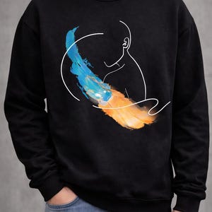 May include: Black sweatshirt featuring a minimalist design. A white outline of a person's head and ear is connected to a blue and orange brushstroke design. The sweatshirt is made of a soft material.