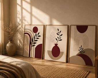 Op de afbeelding: Een set van drie ingelijste wandkunstprints met abstracte ontwerpen in diep bordeauxrood, beige en crème. De illustraties bevatten geometrische bogen, cirkelvormige figuren en botanische bladmotieven met dunne zwarte lijnen.