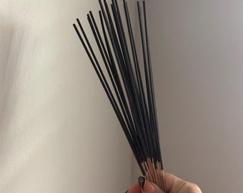 May include: A bundle of thin, black incense sticks with exposed, light brown wooden bases. The sticks are arranged in a fan-like shape, featuring a dark, uniform coating on the upper portions and natural, unfinished wood at the bottom ends.