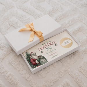 May include: A white rectangular gift box with a gold ribbon bow containing a scratch-off card. The card features a vintage Santa Claus illustration, the text A VERY MERRY CHRISTMAS DAD, A little surprise, just for you, and A ROUND OF GOLF.