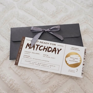 May include: A white rectangular event ticket with brown text reading GET READY FOR MATCHDAY, TICKET HOLDER LEWIS, WEMBLEY STADIUM, LONDON, and a gold scratch-off circle labeled ENGLAND VS SCOTLAND, resting on a dark gray envelope with a gray ribbon.