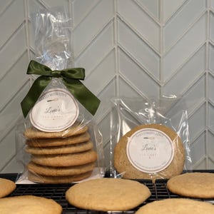 May include: Several golden-brown cookies are displayed. Some are stacked in a clear cellophane bag tied with a green ribbon. The bag has a round label that reads "Baked with Love, Lovie's Tea Cakes." Other cookies are on a black wire rack.