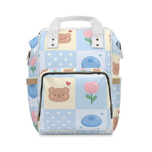 May include: A light blue and white baby backpack with a repeating pattern of cartoon bears, pink flowers, and blueberries. The backpack has a top handle, two side pockets, and a front pocket. The design is a patchwork of squares and polka dots.
