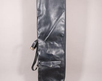 Puede incluir: Una bolsa de viaje negra para palos de golf, con cierre de cremallera y un pequeño bolsillo. La bolsa tiene una correa para el hombro y un ribete rojo en el borde inferior. La bolsa es de material similar al cuero, de unos 130 cm de largo.