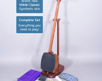 Puede incluir: Un juego completo de shamisen Hibiki Classic con piel sintética. El instrumento de madera se muestra en un soporte, con accesorios que incluyen una tela morada, pequeñas herramientas y una funda con estampado floral. El texto en la imagen dice "Brand-new Hibiki Classic Synthetic skin" y "Complete Set Everything you need to play!"